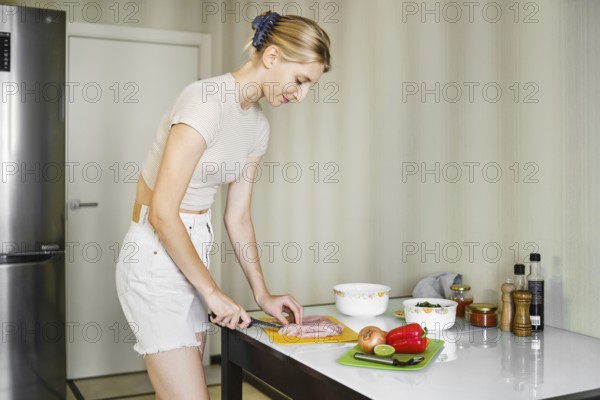 A woman is skillfully chopping chicken on a bright countertop while surrounded by fresh vegetables. The well-lit kitchen features a contemporary design, emphasizing her culinary focus during the day