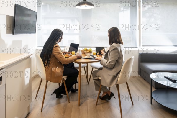 Two businesswomen are enjoying breakfast and working with laptop and tablet in a modern hotel room during their business trip