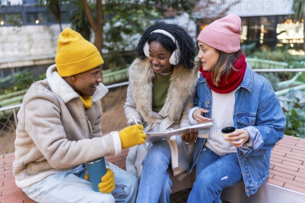 Three happy friends sitting outside in winter, drinking coffee and looking at a map, planning a trip together