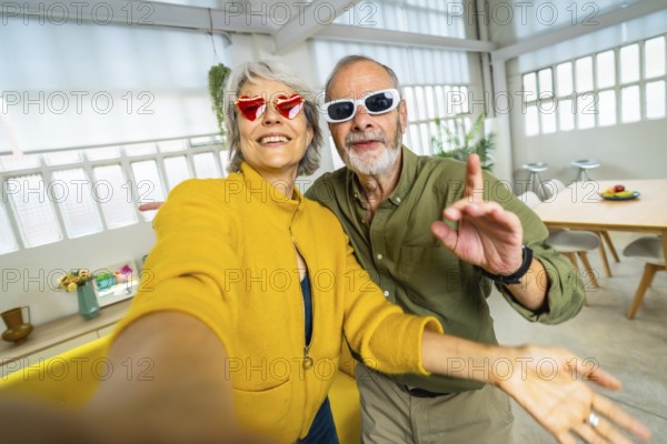 Senior woman with heart shape sunglasses taking selfie with husband while having fun at home