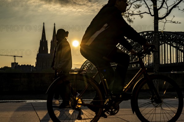 Silhouettes of people in late sunlight, on the Deutz riverbank, right bank of the Rhine, cyclists, on the Rhine promenade, view of Cologne Cathedral and the Hohenzollern railway bridge over the Rhine near Cologne, North Rhine-Westphalia, Germany, Europe