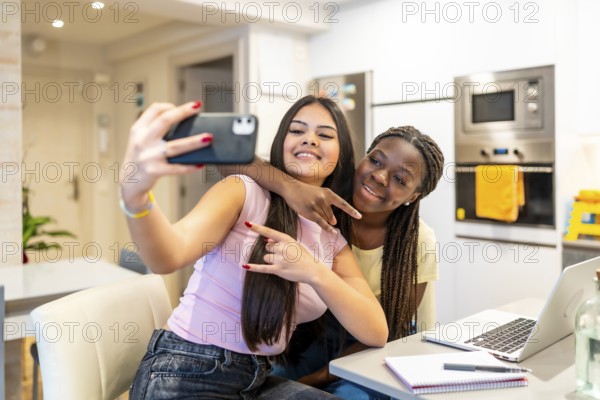 Two happy multi ethnic teenage girls taking a selfie with smartphone while doing homework in a modern kitchen