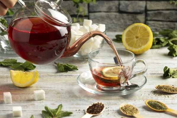 Red tea is poured from a pot into a glass cup with lemon and mint, surrounded by sugar and teas