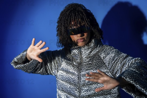 Young woman with curly hair and sunglasses dancing with hands up, wearing a patterned jacket on a vibrant blue backdrop
