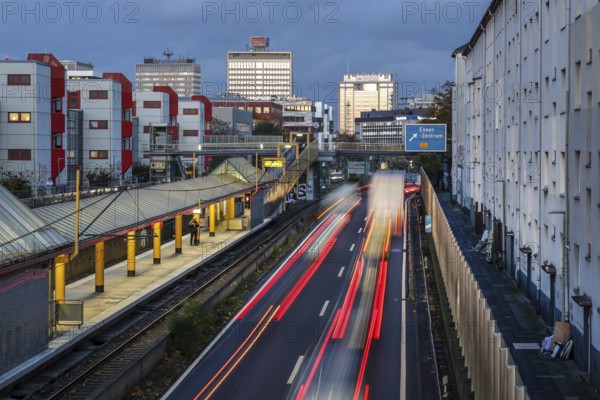Essen, North Rhine-Westphalia, Germany, motorway A40 in the city centre, exit Essen Zentrum, during dusk. Man waiting at Savignystrasse underground station. On the right, residential buildings behind a noise barrier. There is a LANUV air monitoring station, Essen Kruppstraße 117 (EKRU2), on the right under the lantern. Residents are affected by noise and exhaust fumes. Building with Postbank, Eon and Evonik lettering at the back, Europe