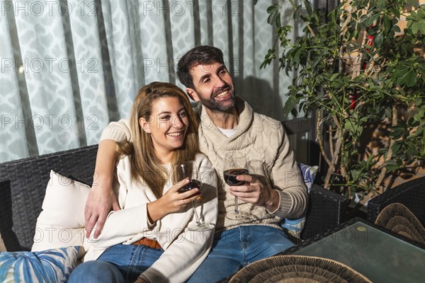 Happy couple relaxing on a comfortable sofa on the terrace, watching a movie and drinking red wine, enjoying their free time together