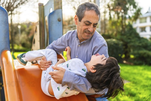 Grandfather catching his grandson sliding down the slide at a playground, having fun together on a sunny day