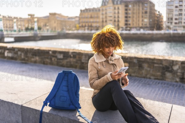 Latin young woman sitting next to the river bank promenade using mobile phone in the city