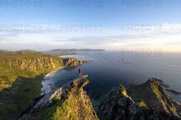 Evening mood, mountaineer standing on a rocky outcrop on the summit of Måtind, view of coastal landscape, Høyvika beach and sea, Måtinden, Andøya island, Vesterålen, Nordland Norway