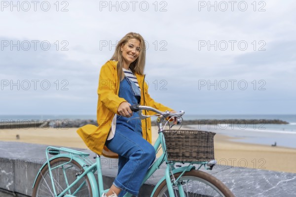 Smiling woman with yellow raincoat sitting on a light blue bicycle by the sea enjoying freedom and healthy lifestyle