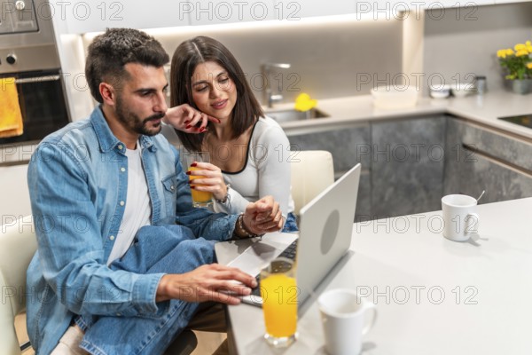 Young couple sitting at kitchen table, drinking orange juice and using laptop, enjoying their time together