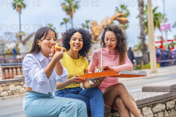 Three female multicultural friends enjoying take away pizza sitting outside the city