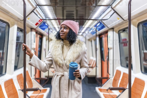 Young black woman wearing winter coat and pink woolen hat holding handrail and reusable coffee cup while riding subway train