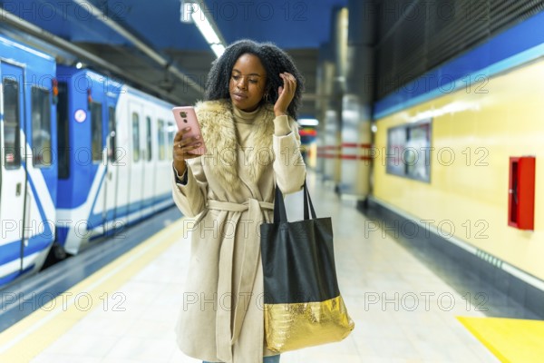Stylish young black woman waiting for subway train arrival while using her mobile phone, staying connected in the urban environment