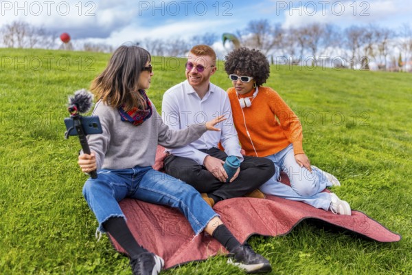 Three content creators are recording an interview for their video podcast in a park, using a smartphone on a selfie stick and an external microphone