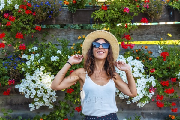 A woman wearing a straw hat and sunglasses is standing in front of a wall of flowers. She is smiling and she is enjoying the beautiful display