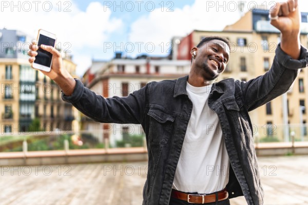 African man dancing happy while listening to music using mobile phone and wireless earbuds in the city