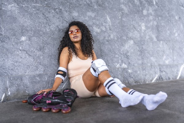 Stylish urban latin woman sitting with inline skates leaning against grey wall in the street