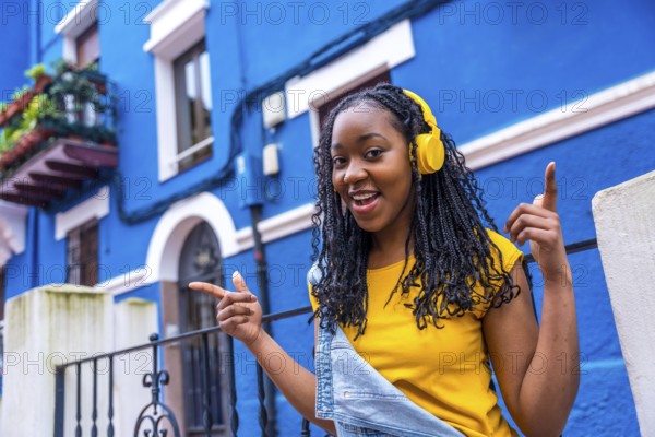 African woman singing while listening to music with headphones standing in a street with blue colored houses