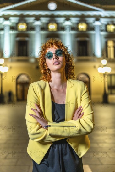 Confident woman in elegant casual clothes and sunglasses standing with arms crossed looking at camera in the city at night