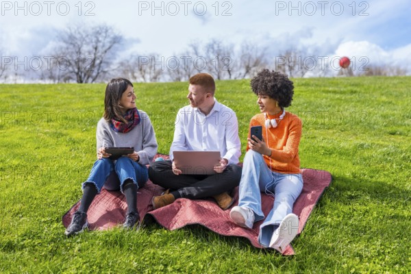 Three university students using electronic devices while sitting on a blanket in a park, enjoying a spring day