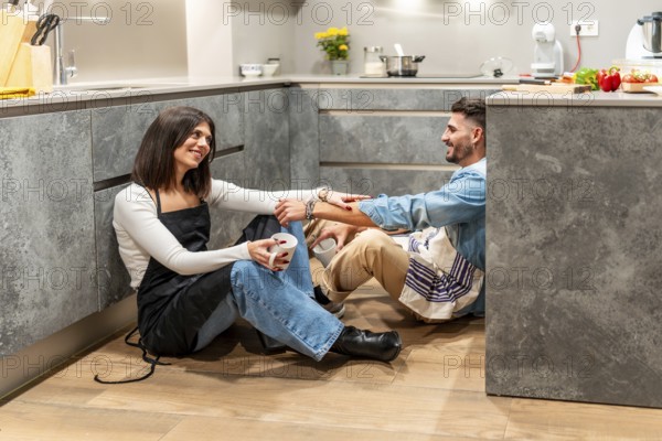 Two chefs are sitting on the floor of their modern kitchen, enjoying a coffee break and chatting after a busy cooking session
