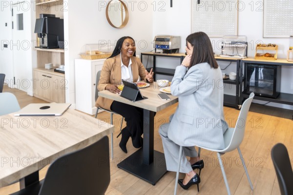 Two businesswomen enjoying breakfast and discussing work during a business trip, utilizing a tablet for their tasks