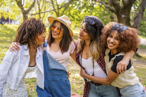 Four diverse young women are enjoying a sunny day in the park, embracing each other and smiling, celebrating their friendship