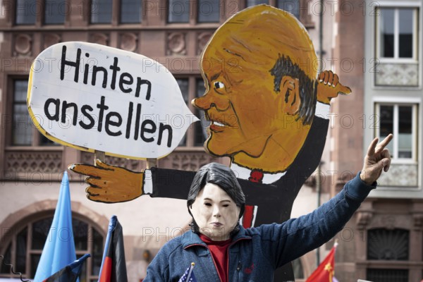 An activist wearing a mask of German Foreign Minister Annalena Baerbock sits on a replica of a tank in front of a speech bubble of German Chancellor Olaf Scholz during the rally for the Easter March on the Römerberg in Frankfurt am Main, Germany, on which it is written Anstellen in the back, meaning climate goals behind rearmament and energy security. A major theme of the rally was the war in Ukraine., Römerberg, Frankfurt am Main, Hesse, Germany, Europe
