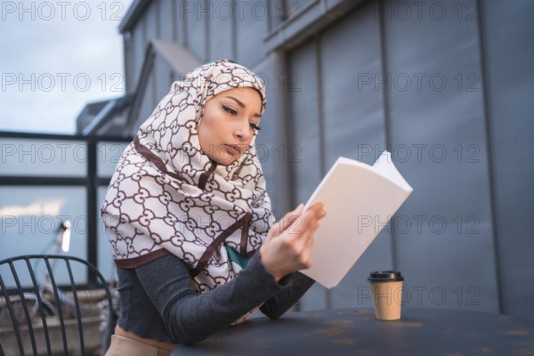 Arab girl with white veil at the computer on the terrace of a coffe shop, having a lot of fun reading a beautiful book