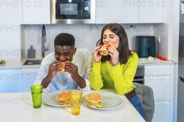 Young multiethnic couple eating homemade burgers in their modern kitchen, enjoying a casual meal together