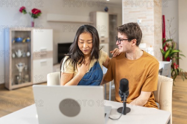 Asian woman and caucasian man recording a podcast at home, showcasing a modern lifestyle with technology and casual interaction