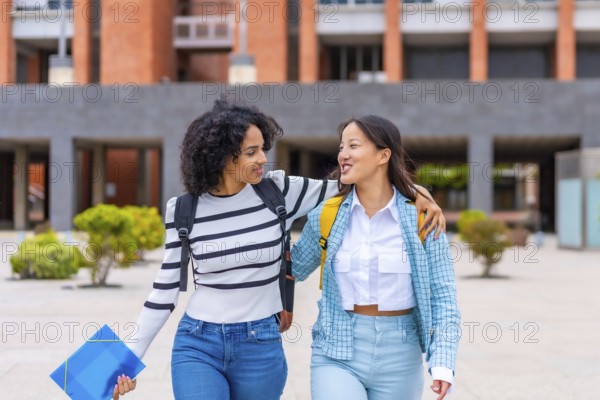 Frontal three quarter length photo of two multi-ethnic female student embracing walking along the university campus