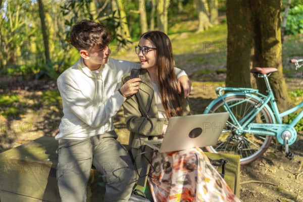 Two students are working on a laptop in a park, charging it with a power bank while sitting on a bench near their bicycle