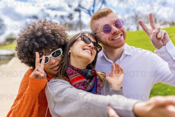 Three happy multi ethnic friends wearing sunglasses gesturing peace sign while taking a selfie in a park