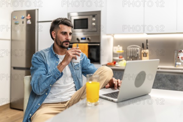 Man savoring a cup of coffee while working remotely on a laptop in a stylish, modern kitchen, enjoying a productive morning routine