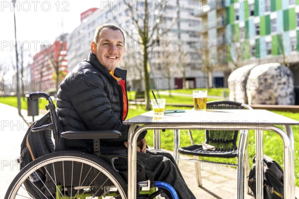 Young man in a wheelchair enjoys a refreshing drink with a friend at a table outside a cafe, embracing the social atmosphere and urban surroundings