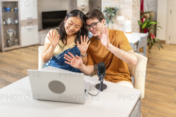Multiethnic couple of content creators waving at their audience while recording a new episode of their podcast from home