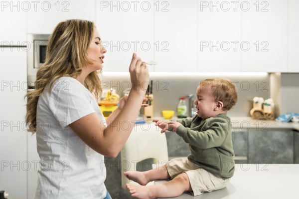 Young mother struggling to feed her crying baby boy while sitting on the kitchen counter at home, navigating the challenges of mealtime