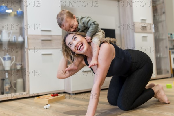 Young mother joyfully playing with her baby son, carrying him on her back while enjoying quality time together in the living room