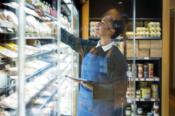 Smiling saleswoman wearing blue apron checking food products using digital tablet while working in organic supermarket