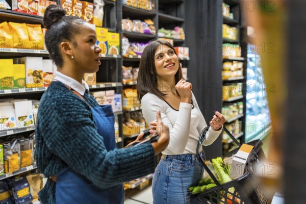 Saleswoman using tablet, advising customer holding shopping basket, choosing products on shelves in organic supermarket
