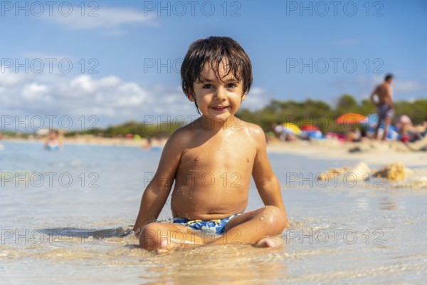 A young boy is sitting in the water with his arms crossed. He is smiling and he is enjoying himself