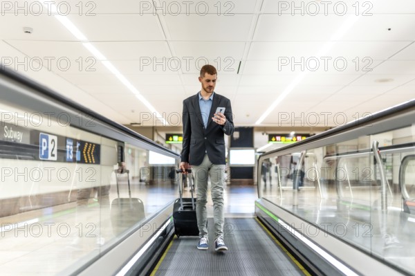 Businessman pulling suitcase and using smartphone while walking on moving walkway at airport