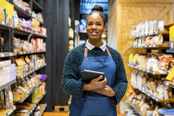 Confident saleswoman in an apron, smiling while holding a digital tablet and working among shelves of organic products