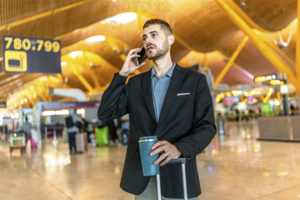 Businessman waiting for his flight, talking on the phone and holding a coffee while pulling his suitcase in a modern airport