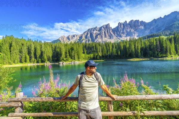 Hiker relaxing by a wooden fence, admiring the stunning turquoise waters of lake carezza with the latemar massif in the background, dolomites, italy
