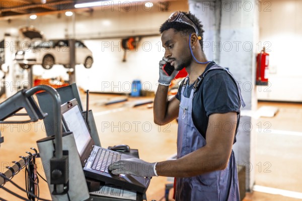 Young mechanic using a laptop and talking on the phone while working on a car in a professional workshop