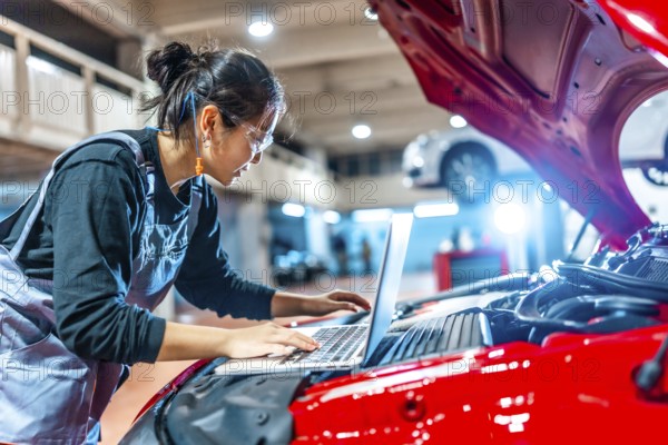 Young female mechanic using a laptop while inspecting a car engine in a professional auto repair shop
