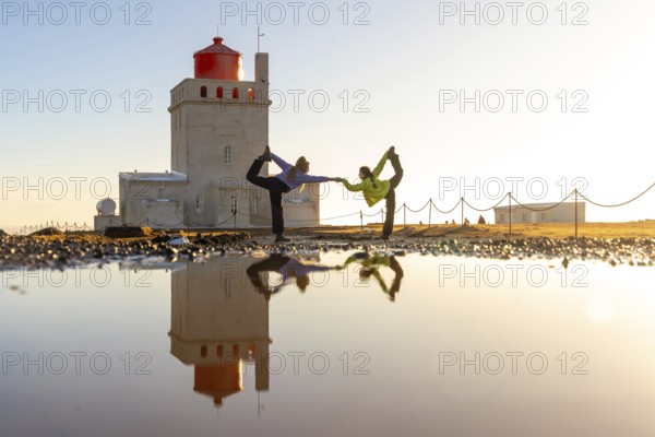 Two people are doing yoga on a beach in front of a lighthouse. The water is calm and the sky is clear
